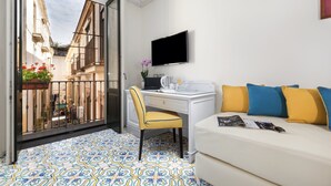 Une pièce avec un sol à motifs, un bureau avec une chaise, un téléviseur accroché au mur, un canapé avec des coussins et une vue sur la rue, visible à travers une porte vitrée.