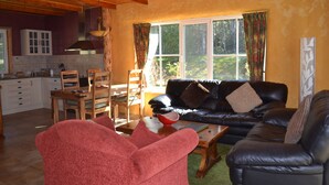 A cozy living room with a black leather sofa, red armchairs, and a wooden coffee table. A kitchen with white cabinets and a dining area with wooden chairs and a table are visible in the background. - Whispering Spirit Holiday Cottages (Franklin)