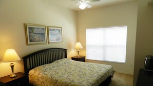 A bedroom with a bed, two bedside lamps, a ceiling fan, a window with blinds, and two framed pictures on the wall.