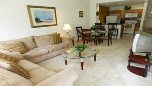 A living room with a beige sofa, a glass coffee table, and a dining area with wooden chairs and a glass table. A kitchen with wooden cabinets and a microwave is visible in the background.
