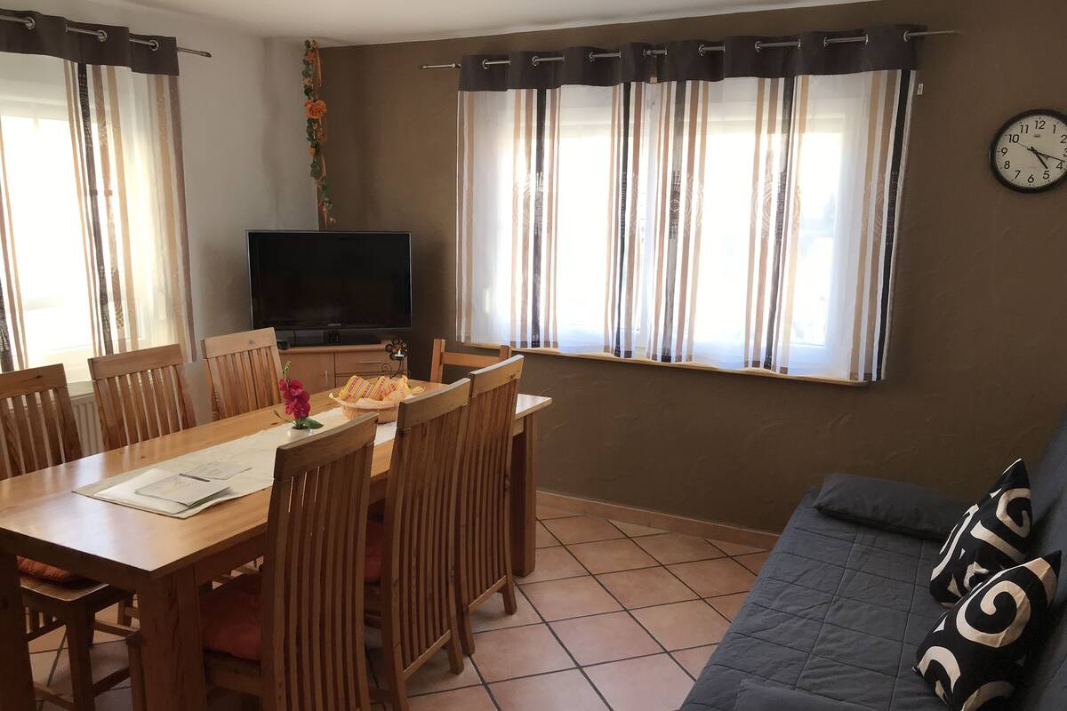 A living room with a dining table, chairs, a sofa, a TV, and windows with curtains.