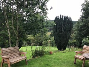 Eine hölzerne Bank in einem üppigen Garten mit einem hohen Baum und Blick auf ein entferntes Feld.