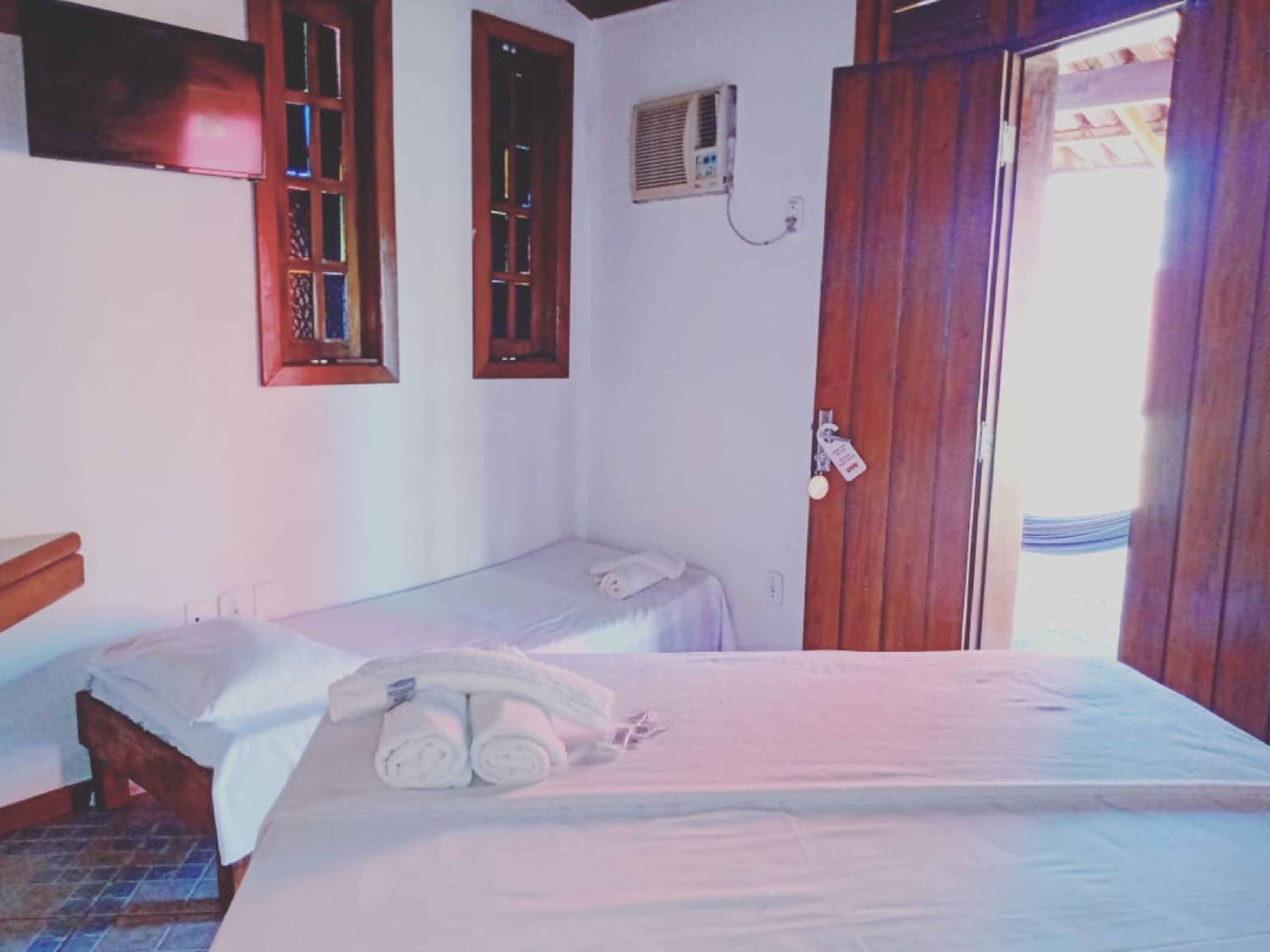 A room with two single beds, a wooden door, a wall-mounted air conditioner, and a television on the wall.