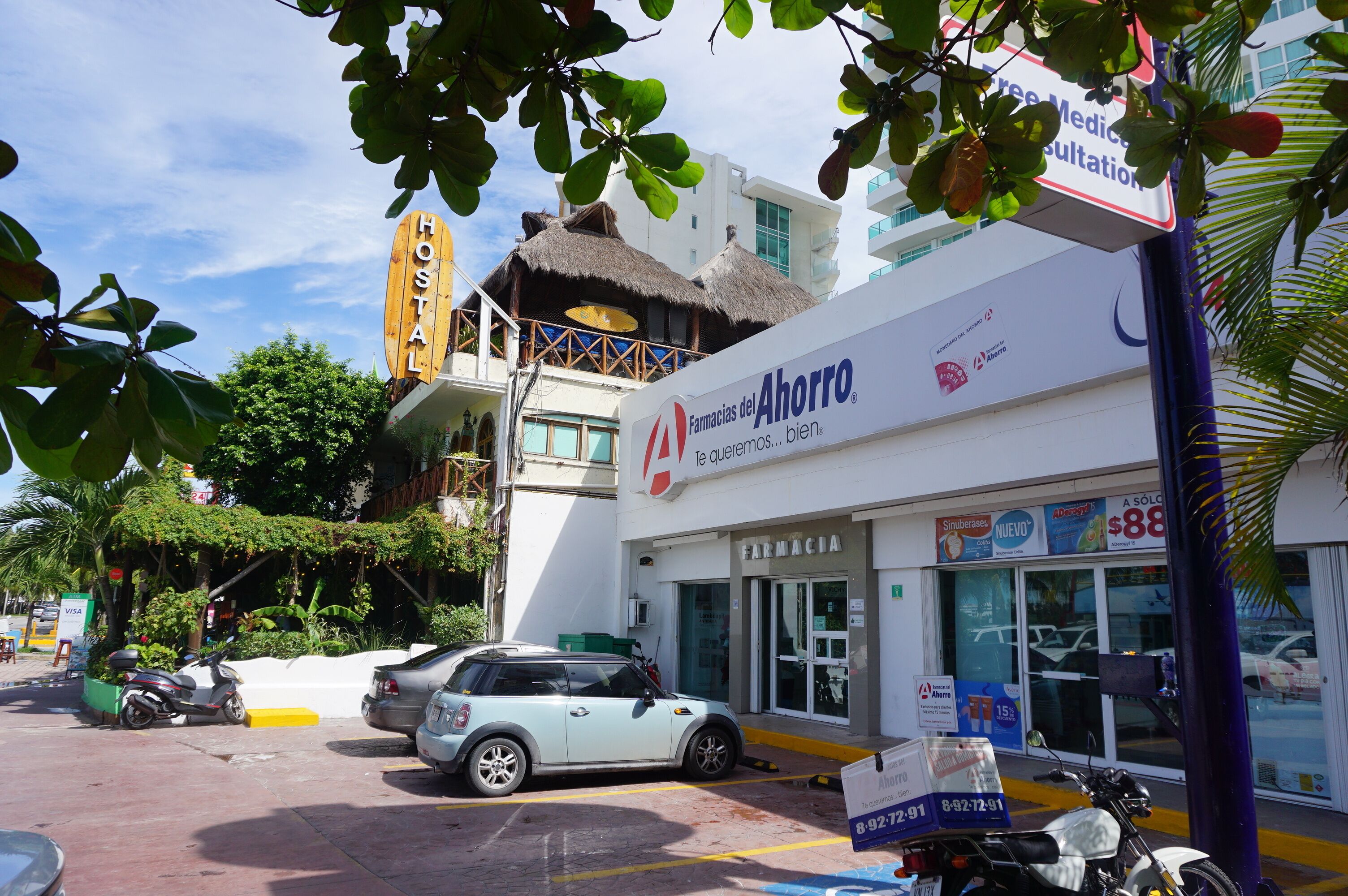 Una farmacia llamada 'Farmacias del Ahorro' con un letrero, un coche blanco estacionado al frente y una estructura de techo de paja al fondo.