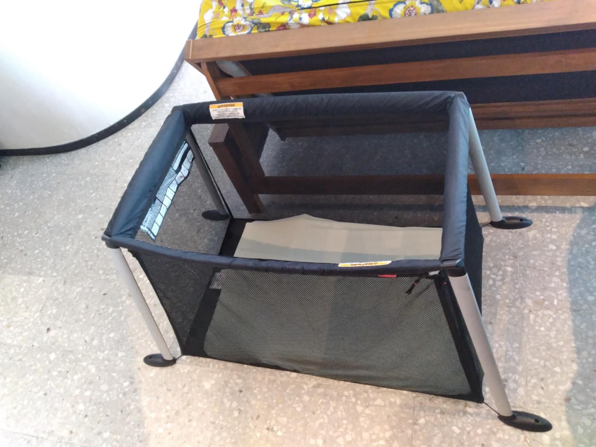 Una cuna portátil para bebé con parte superior de malla y base de madera, colocada sobre un suelo de baldosas.