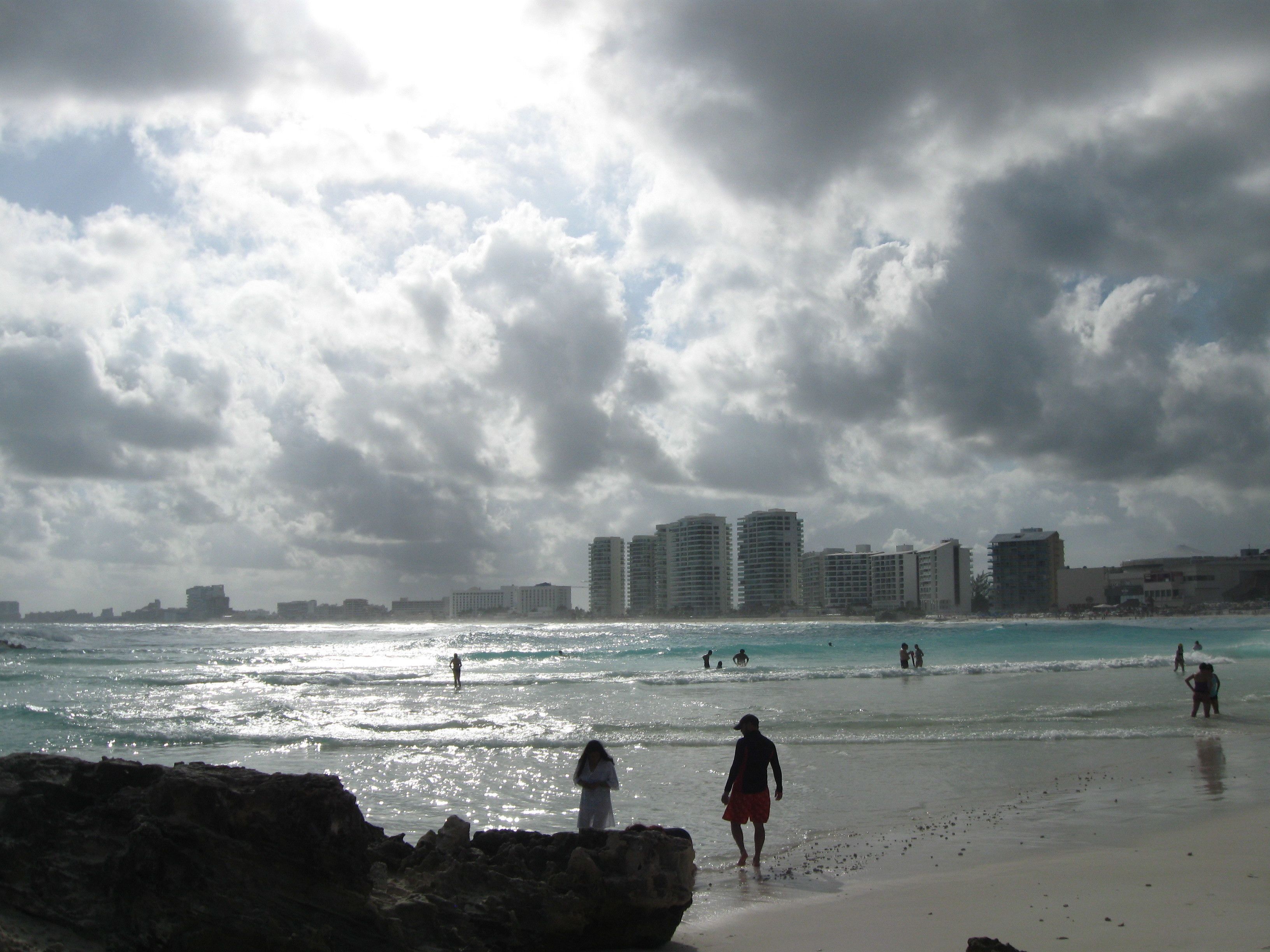 A beach with people walking along the shore, a rocky foreground, and buildings in the background.