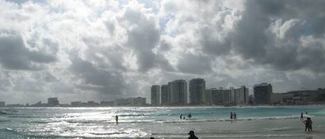 A beach with people walking along the shore, a rocky foreground, and buildings in the background.