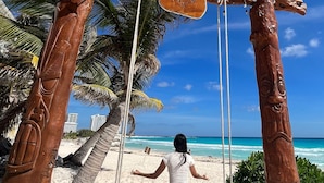 A wooden swing set with a woman sitting on it, overlooking a beach with palm trees and the ocean. - Ocean Dream 1 Bedroom Condo (Cancun)