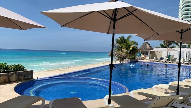 A pool area with lounge chairs and umbrellas, a beach view, and a thatched-roof structure. - Ocean Dream 1 Bedroom Condo (Cancun)