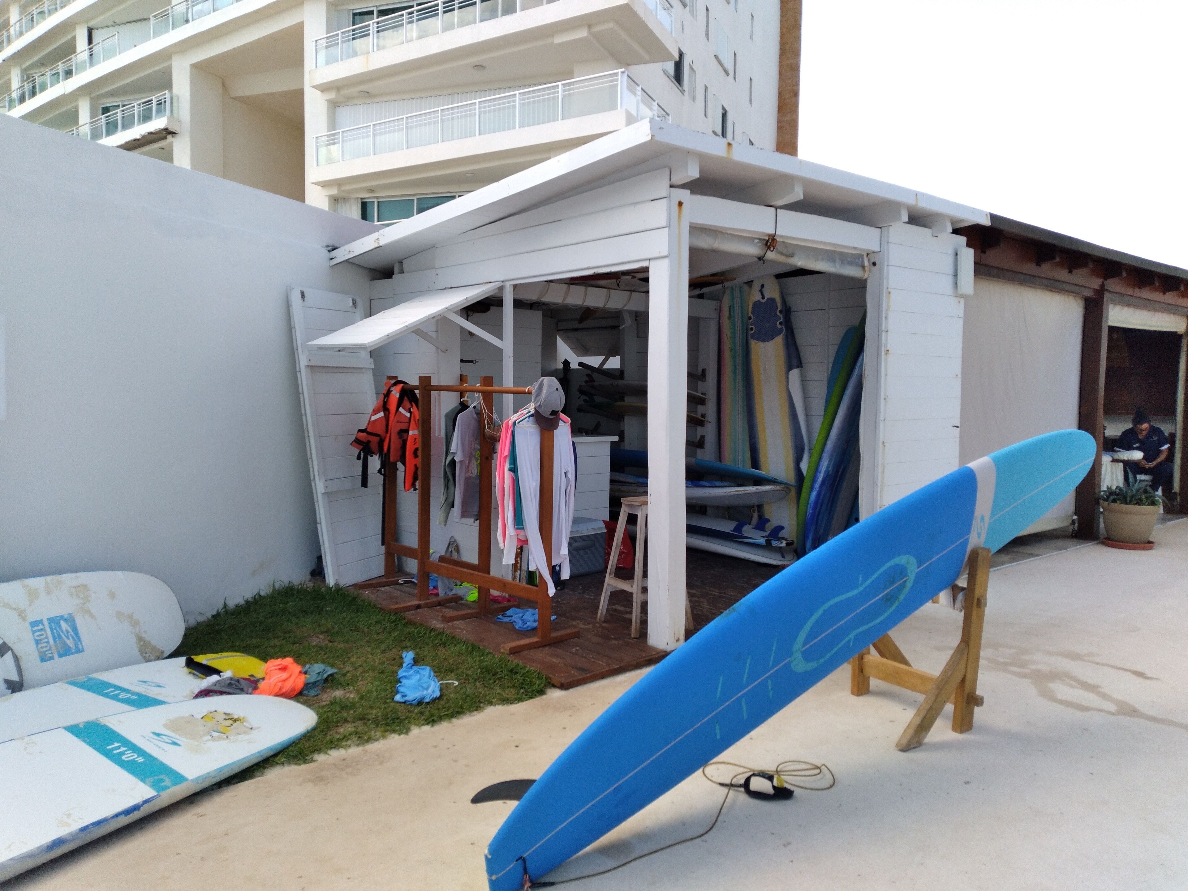 A building with a white exterior, a small outdoor area with a surfboard on a stand, and a clothing rack with various items.