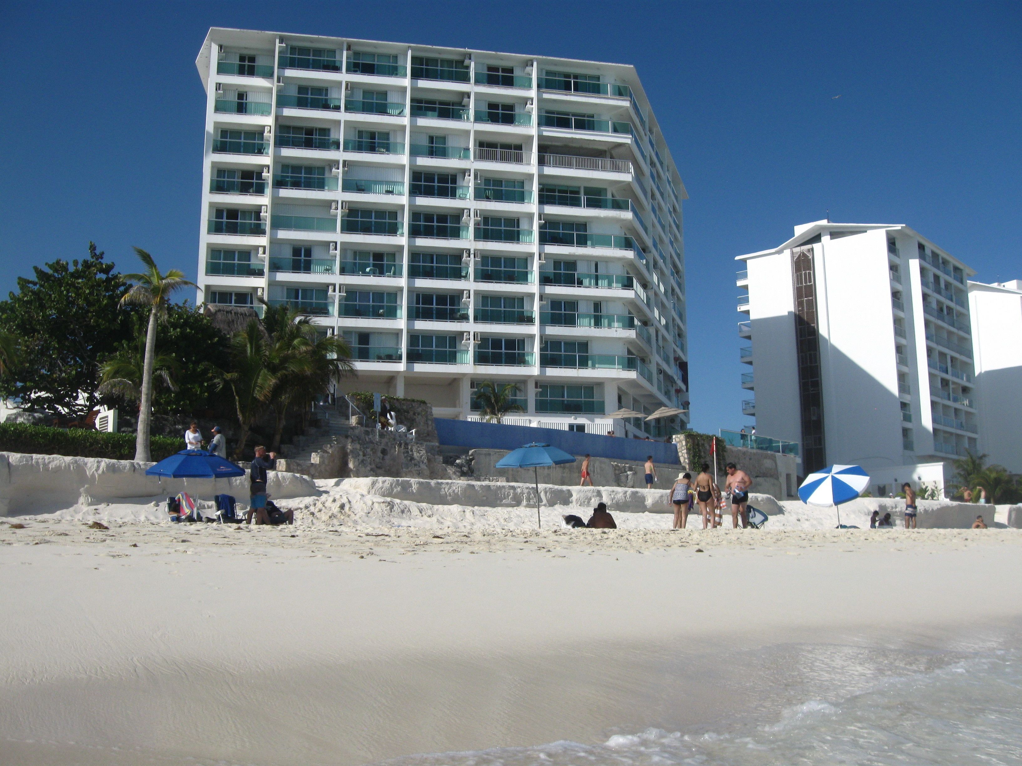 Un hôtel situé en bord de mer, avec plusieurs étages, des balcons et une plage de sable équipée de parasols.