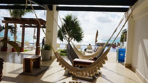 A hammock chair with a view of the ocean, a wooden table, and potted plants in a sunny outdoor area. - Ocean Dream 1 Bedroom Condo (Cancun)
