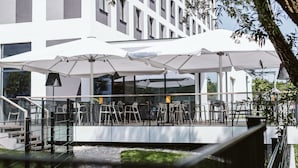 Een terras buiten met parasols, tafels en stoelen, met uitzicht op een waterpartij en een modern gebouw.