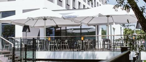 An outdoor seating area with umbrellas, tables, and chairs, overlooking a body of water and a modern building.