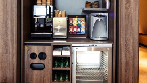 Una kitchenette compacta con cafetera, tostadora, mini refrigerador y estantes con tazas y vasos.