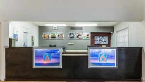 A reception desk with two monitors displaying the '6 Extended Stay' logo, a bulletin board with various notices, and a wall-mounted television. - Studio 6 Alexandria, LA (Alexandria)