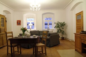 A dining area with a wooden table, chairs, and a vase of flowers. There is a sofa and a potted plant in the background. - BioRezydencja (Swieradow-Zdroj)