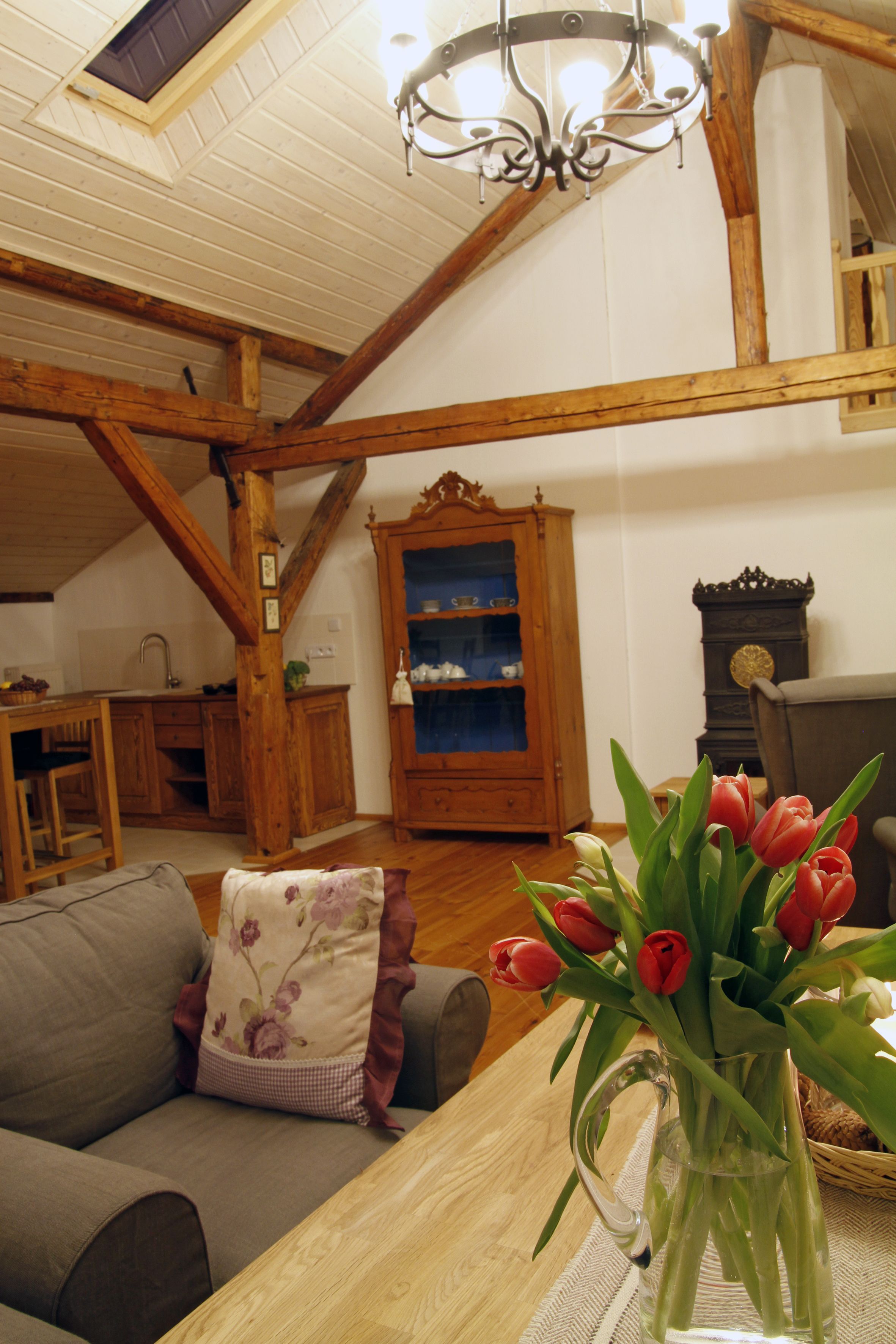 Een gezellige woonkamer met een houten plafond, een kroonluchter, een bank met bloemenkussens en een vaas rode tulpen op een houten tafel.