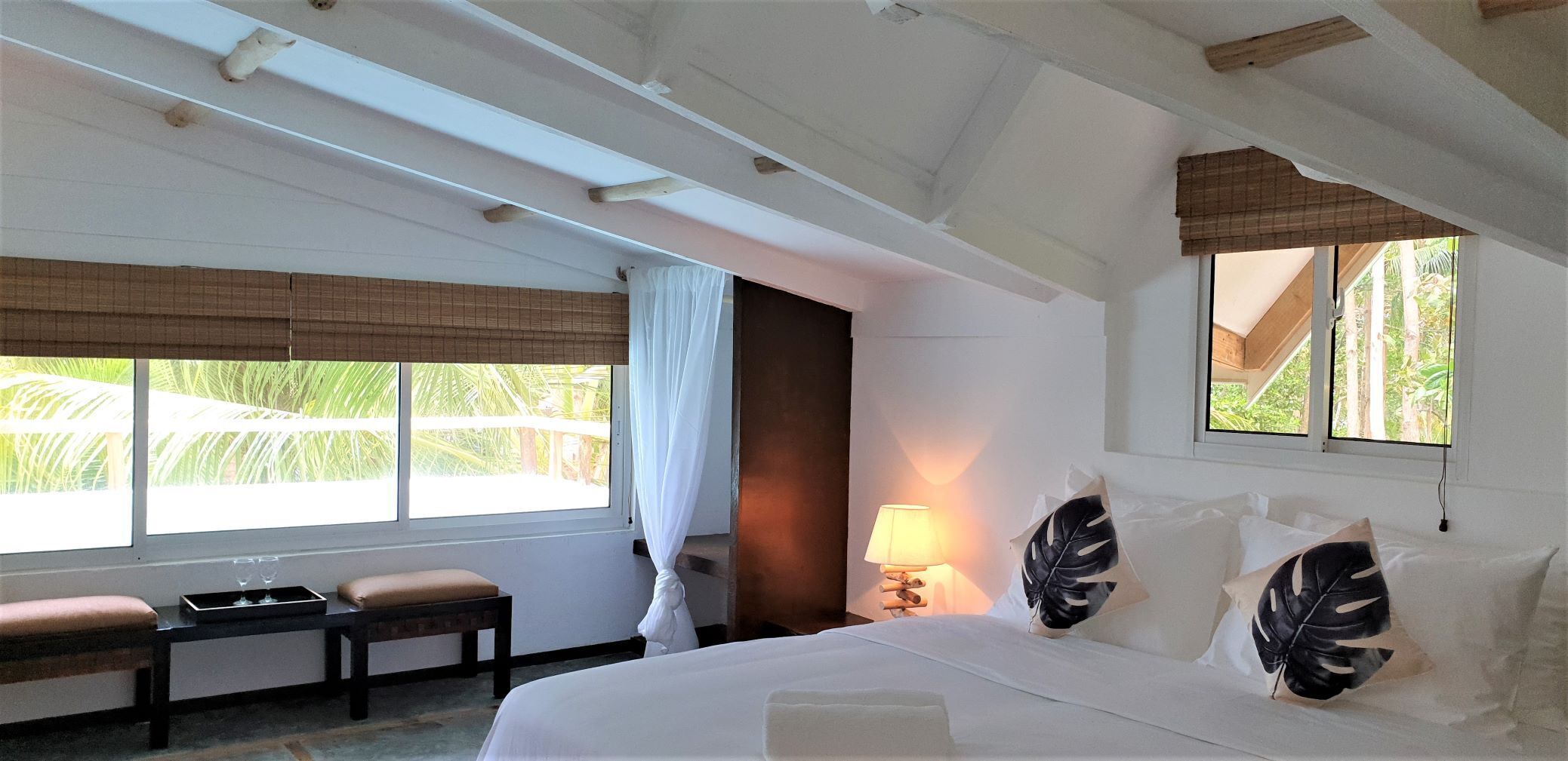 A hotel room with a bed, bedside table, lamp, and a view of palm trees through the window.