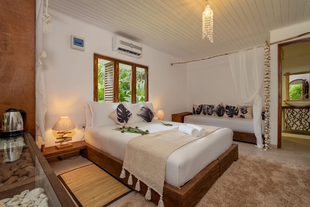 Quarto de hotel com uma cama grande, uma mesa de cabeceira com chaleira, abajur e vista para o verde pela janela.