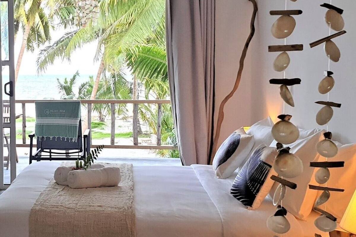 Quarto com cama, almofadas, mesa de cabeceira e vista para a praia.