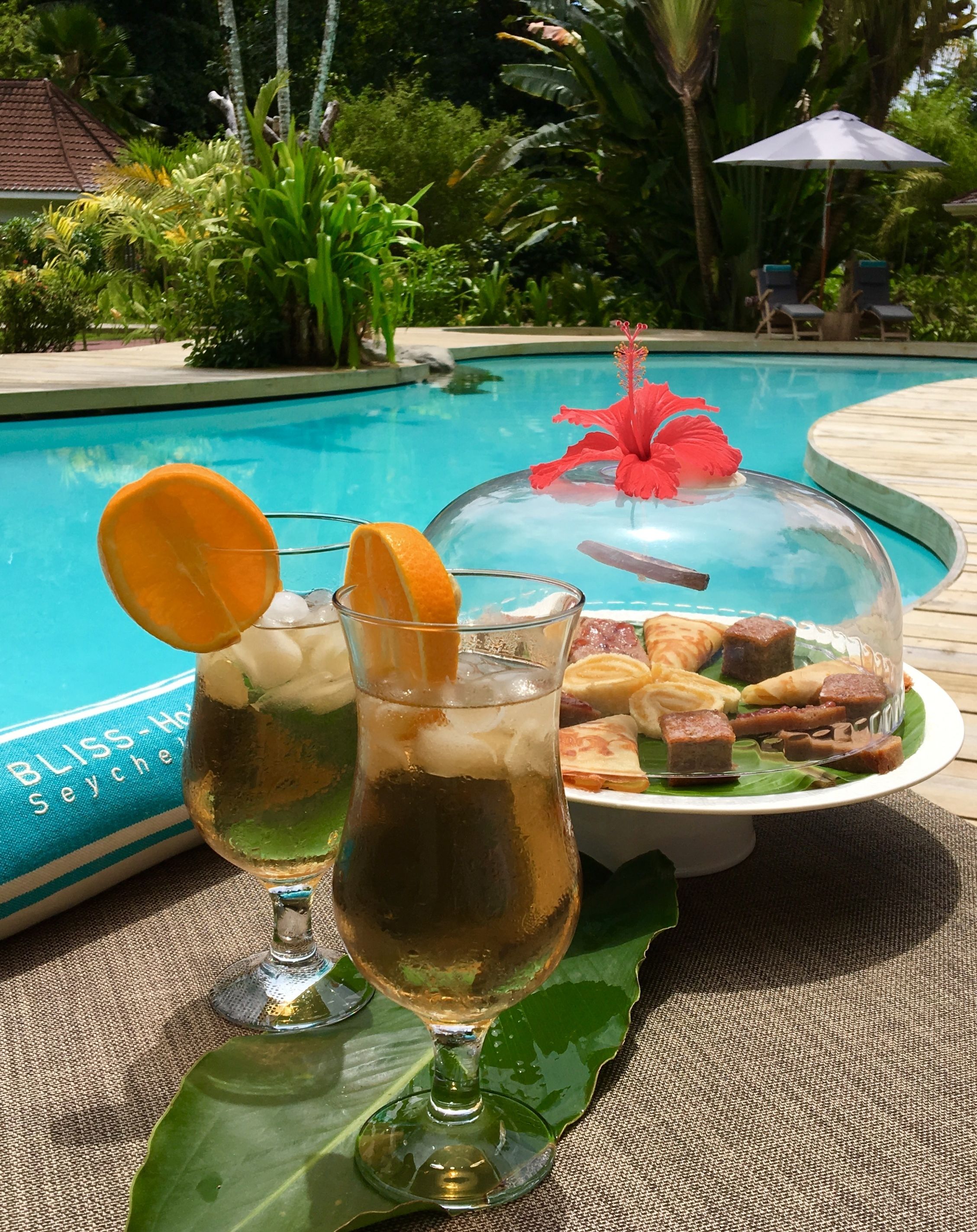 A poolside setting with a glass of iced beverage, a covered food display, and a tropical environment.