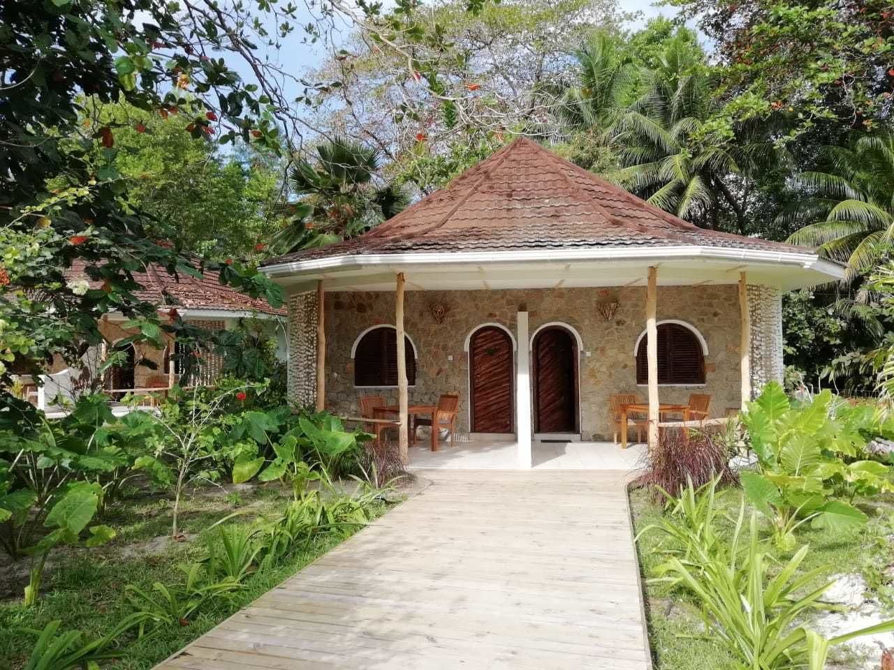 A small, single-story building with a tiled roof and three arched doorways. It is surrounded by lush greenery and has a paved pathway leading to the entrance.