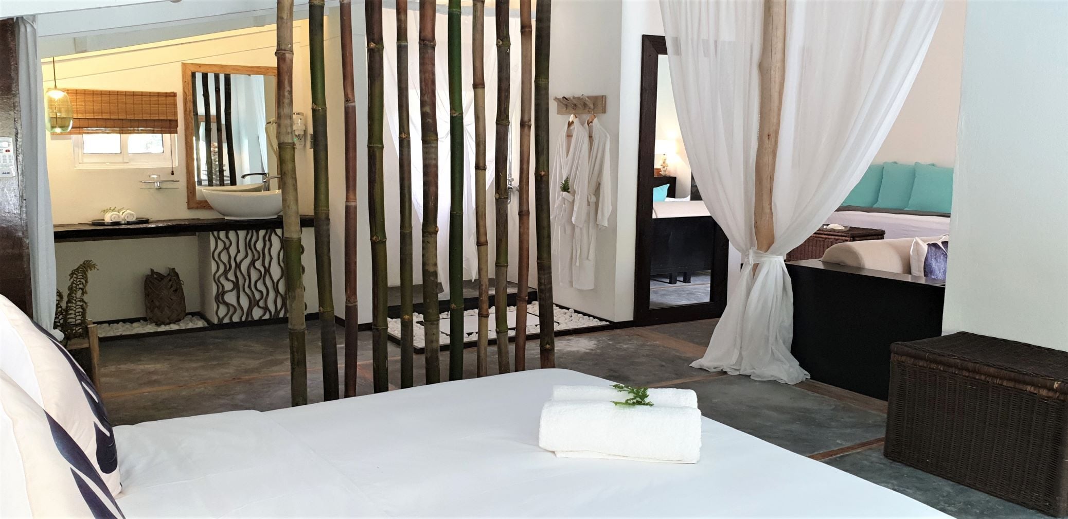 Une chambre à coucher avec un lit, une cloison en bois, une salle de bain visible à travers une porte en verre et un rideau blanc.