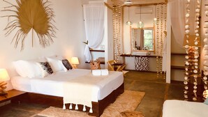 A bedroom with a bed, bedside tables, a nightstand, a wooden stool, and a bathroom with a sink and mirror. - Bliss Hotel Praslin (Praslin Island)