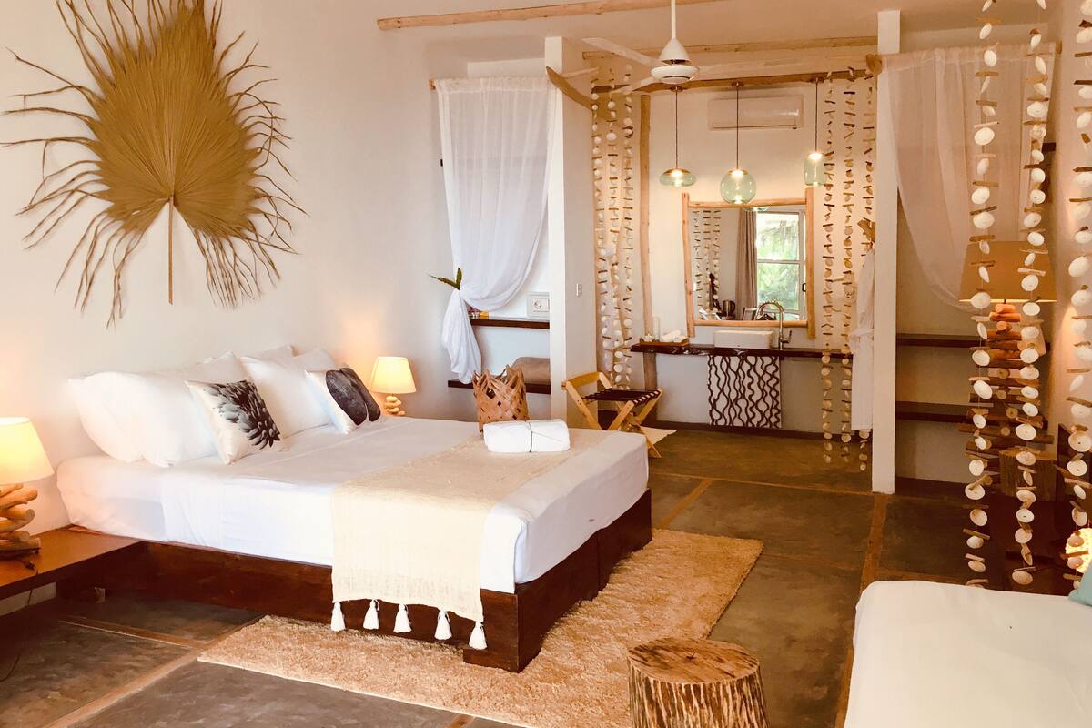 Quarto com cama, mesas de cabeceira, um banquinho de madeira e banheiro com pia e espelho.