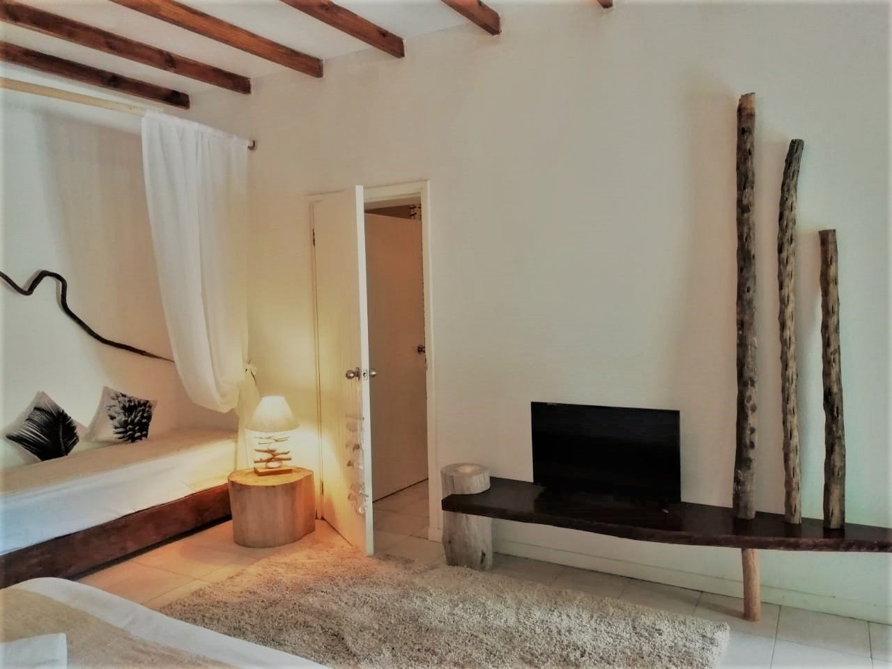 A bedroom with a bed, a TV on a wooden stand, a door leading to another room, and a small table with a lamp.