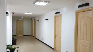 Un pasillo largo y estrecho con suelo de baldosas color beige y paredes blancas. Se observan varias puertas a la derecha, cada una con un número. El pasillo está bien iluminado con luces de techo.