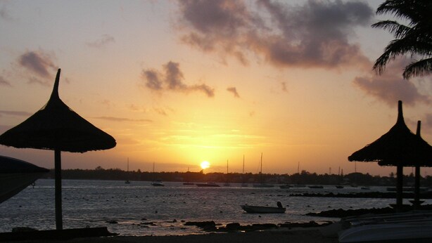 Escena de playa con dos sombrillas de paja y un atardecer sobre el agua.