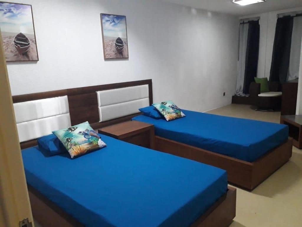 Ein Zimmer mit zwei Betten, jeweils mit blauer Matratze und passendem Kissen, einem Holzkopfstück und einem kleinen Tisch mit Stuhl.