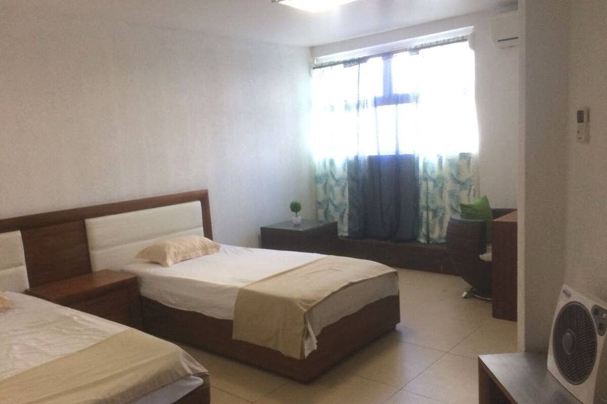 Una habitación de hotel con dos camas, una mesita, una ventana con cortinas y un aire acondicionado instalado en la pared.