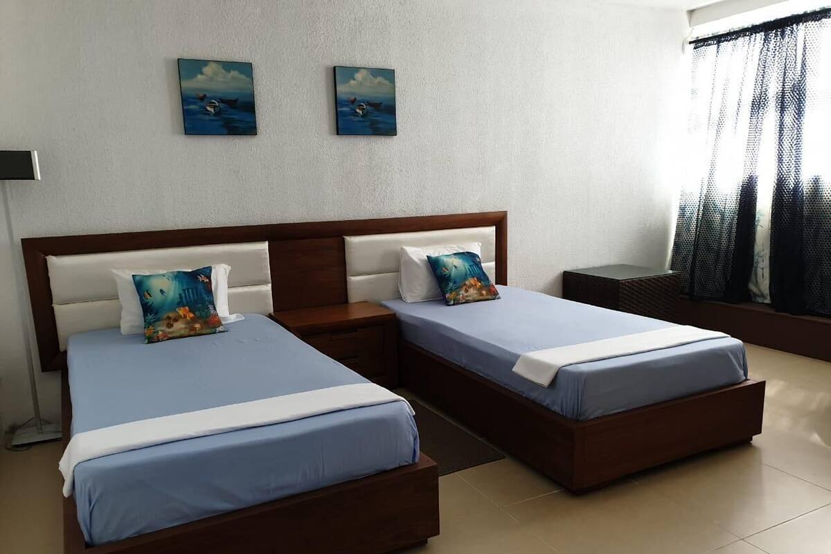 Habitación de hotel con dos camas individuales, cabecera de madera, mesita de noche y un ventanal con cortinas translúcidas.