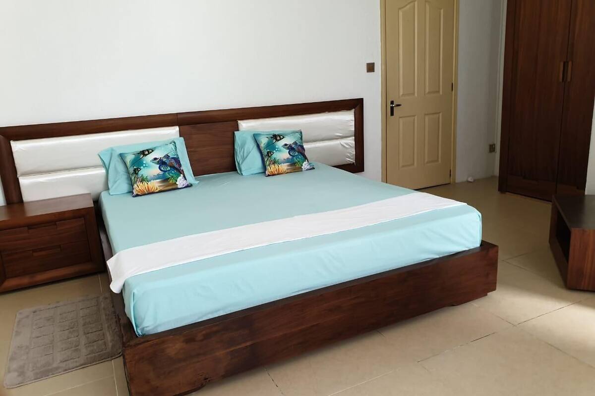 Una cama con estructura de madera y cubrecama azul claro, dos almohadas a juego y una mesita de noche de madera.