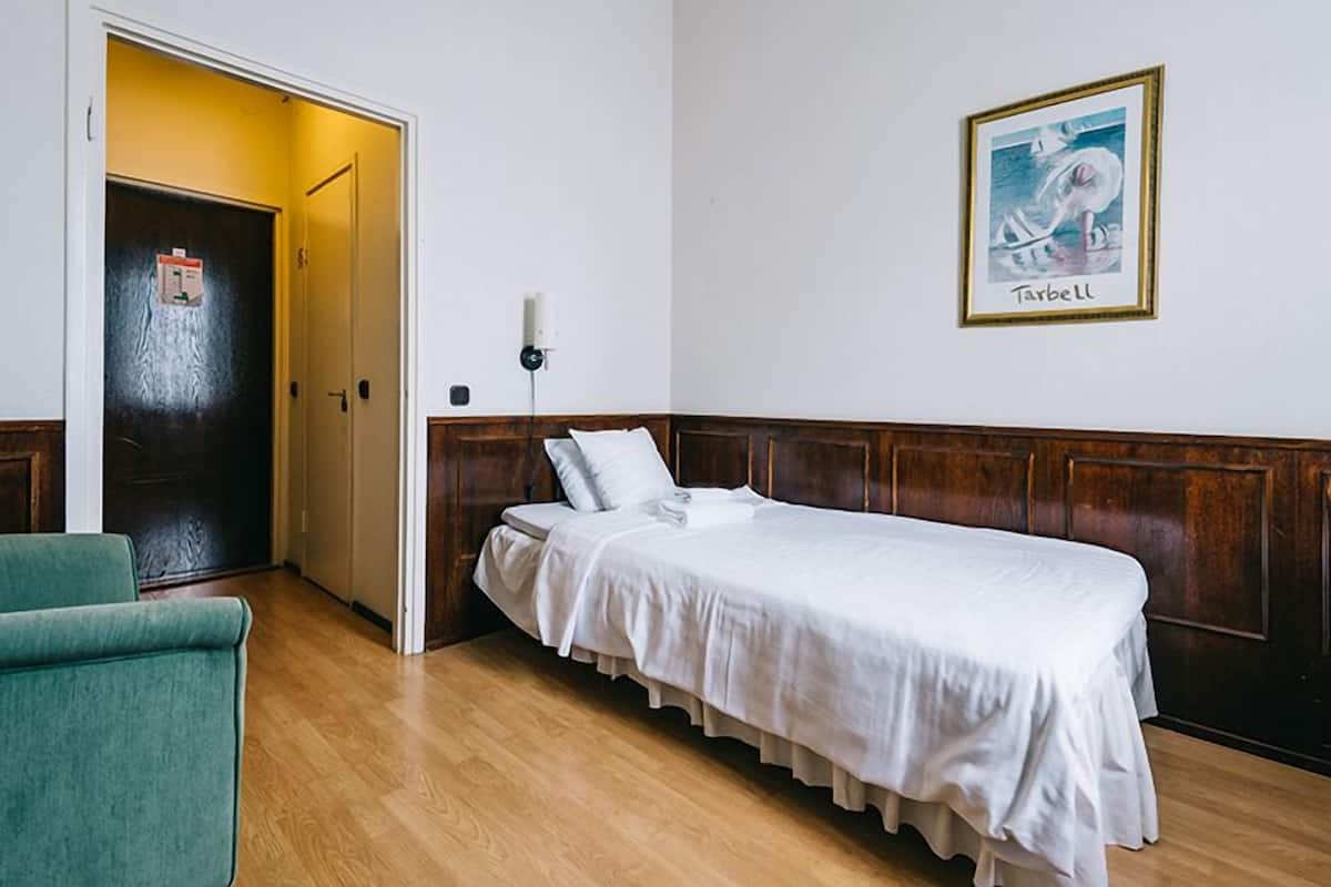 A room with a single bed, a green chair, a door with a fire alarm, and a framed picture on the wall.