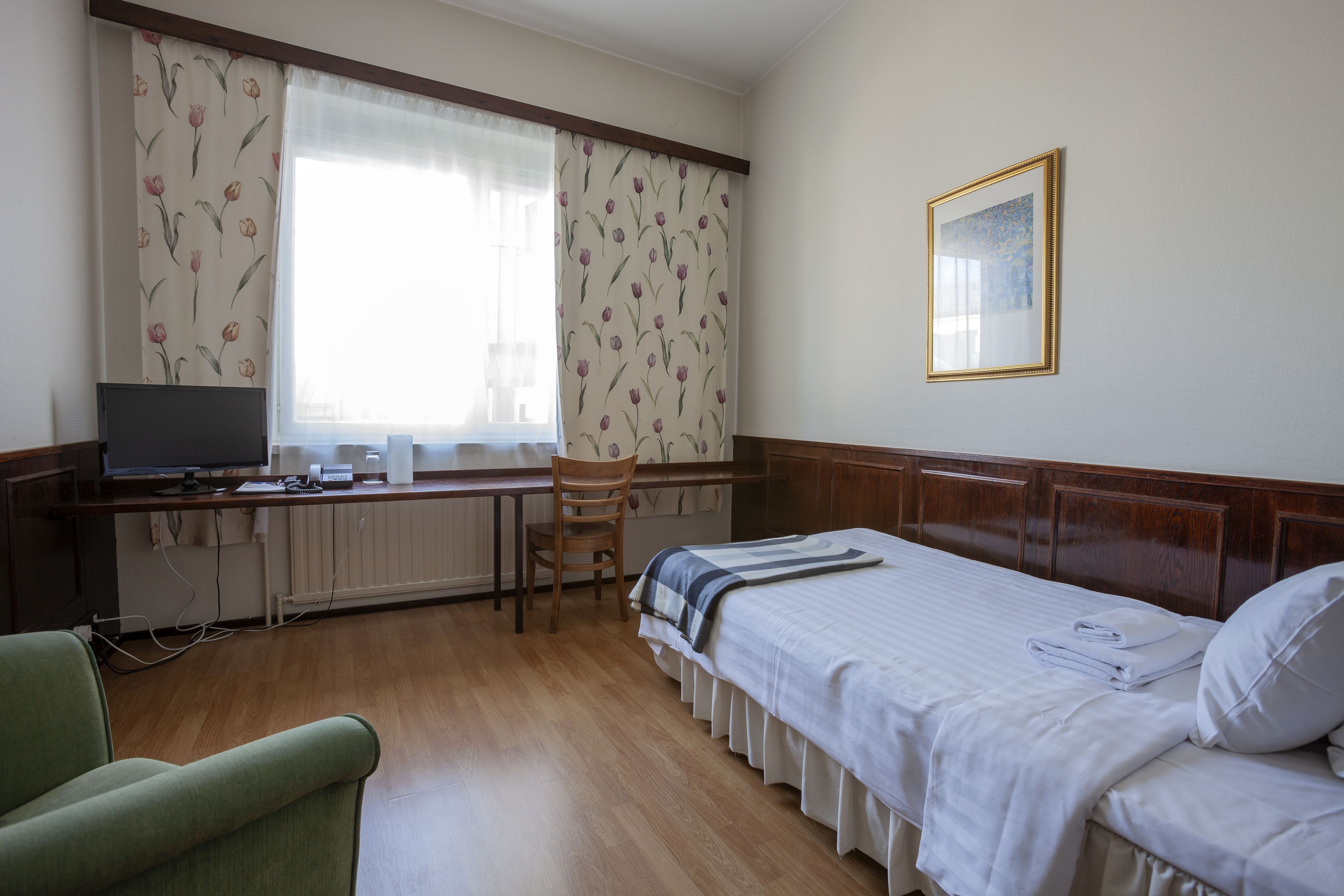 A hotel room with a bed, a desk with a computer, a chair, and a window with curtains.