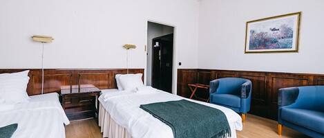A hotel room with two beds, a wooden headboard, a blue armchair, a small table, and a framed picture on the wall.