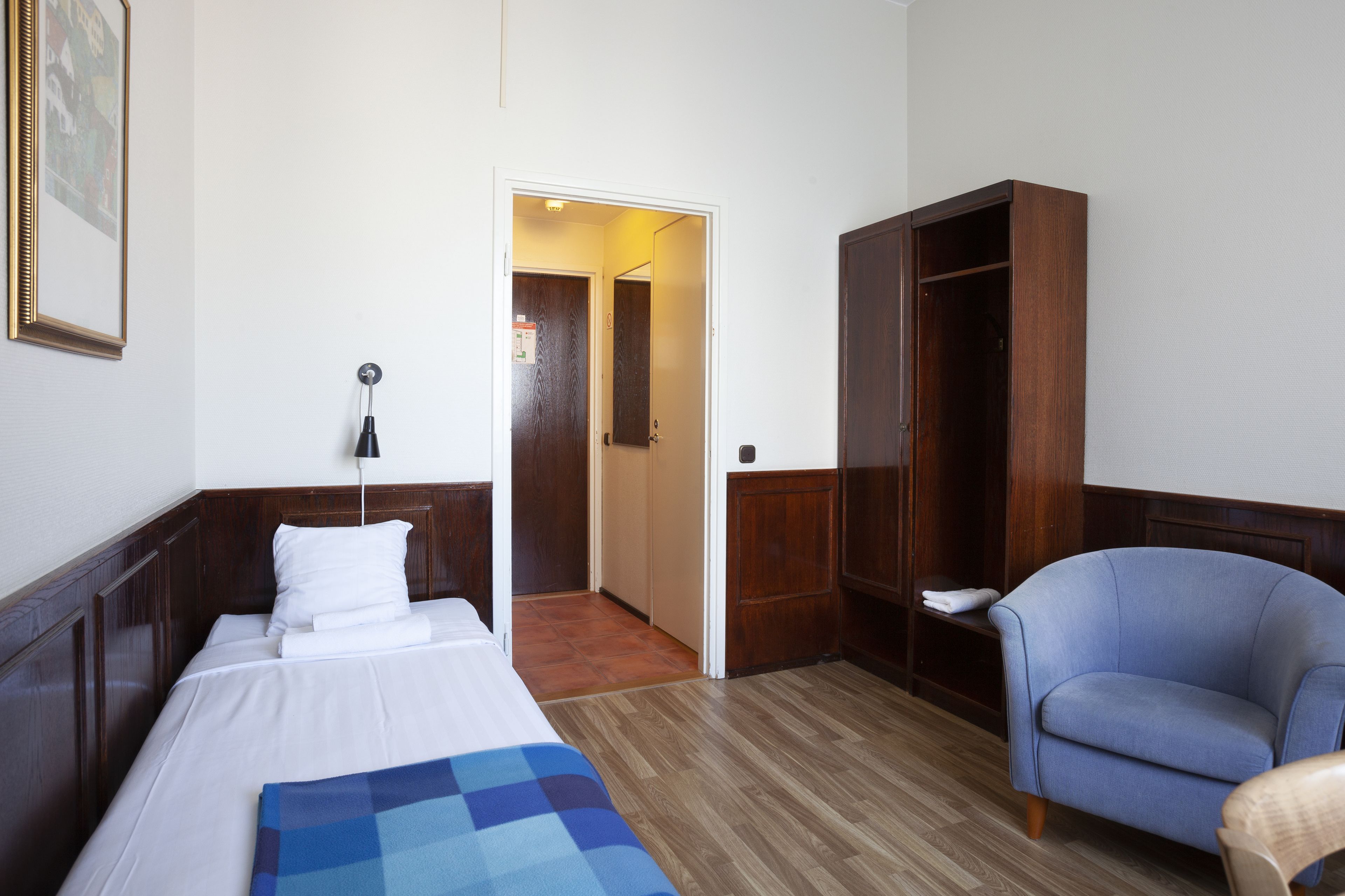 Una habitación de hotel con una cama, un sillón azul, un armario de madera y una puerta que da a otra habitación.