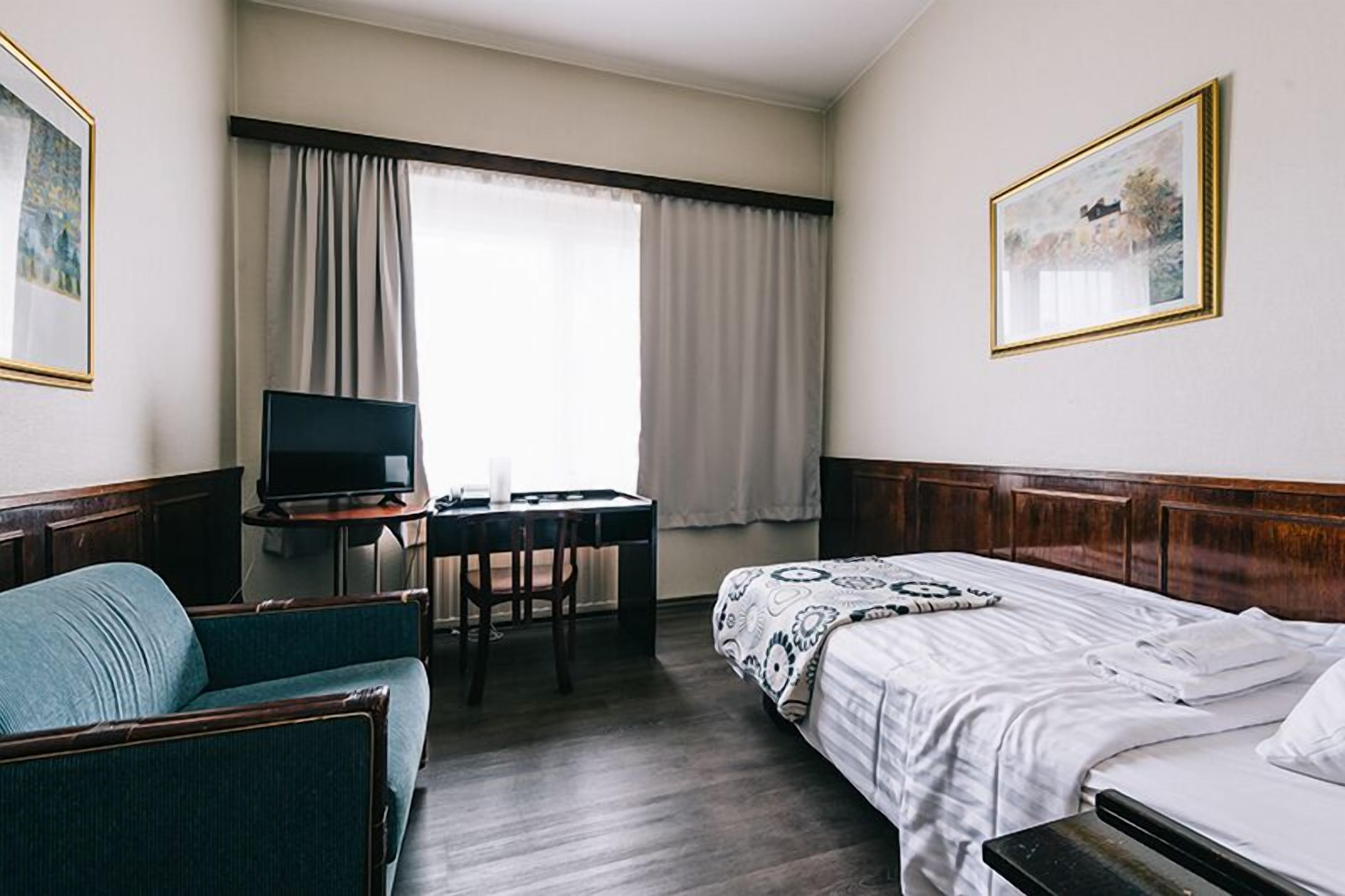 A hotel room with a bed, a desk with a chair, a television, and a window with curtains.