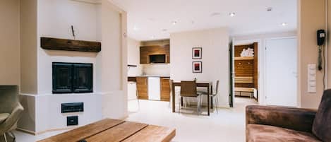 A modern living room with a wooden coffee table, a brown sofa, and a TV mounted on the wall. There is a dining area with a table and chairs, and a kitchen in the background.