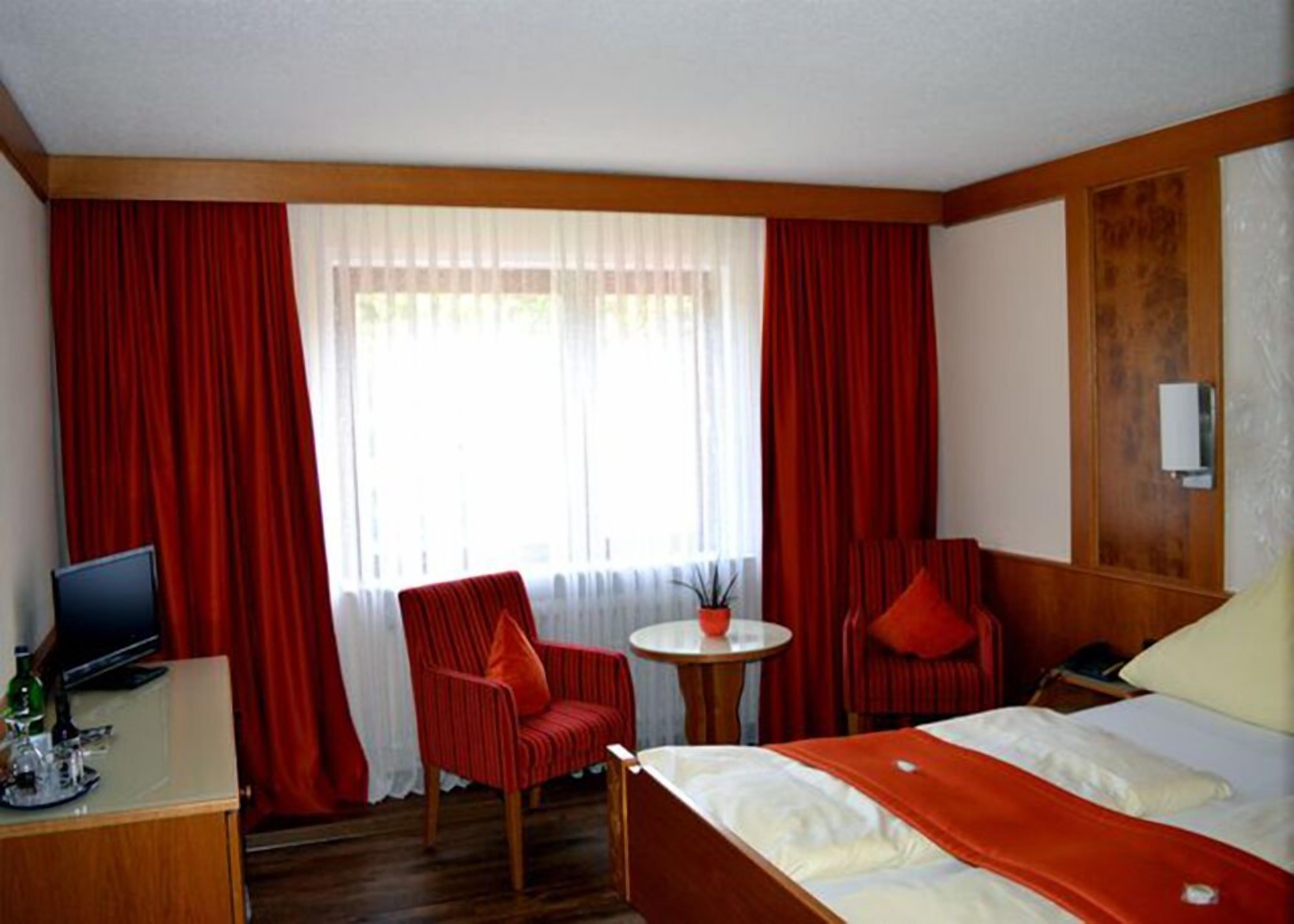 Et hotelværelse med en seng, et skrivebord med computer, en stol, et lille bord og et vindue med gardiner.
