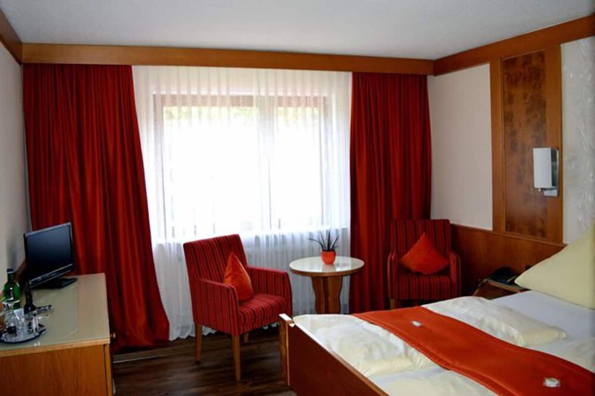 Une chambre d’hôtel comprenant un lit, un bureau avec un ordinateur, une chaise, une petite table et une fenêtre avec des rideaux.