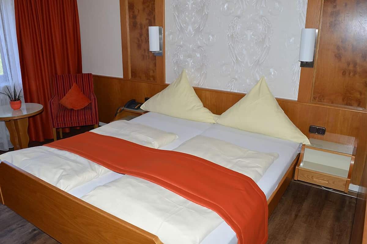 A hotel room with a large bed, a wooden headboard, a small table with a plant, a chair, and a nightstand with a lamp.