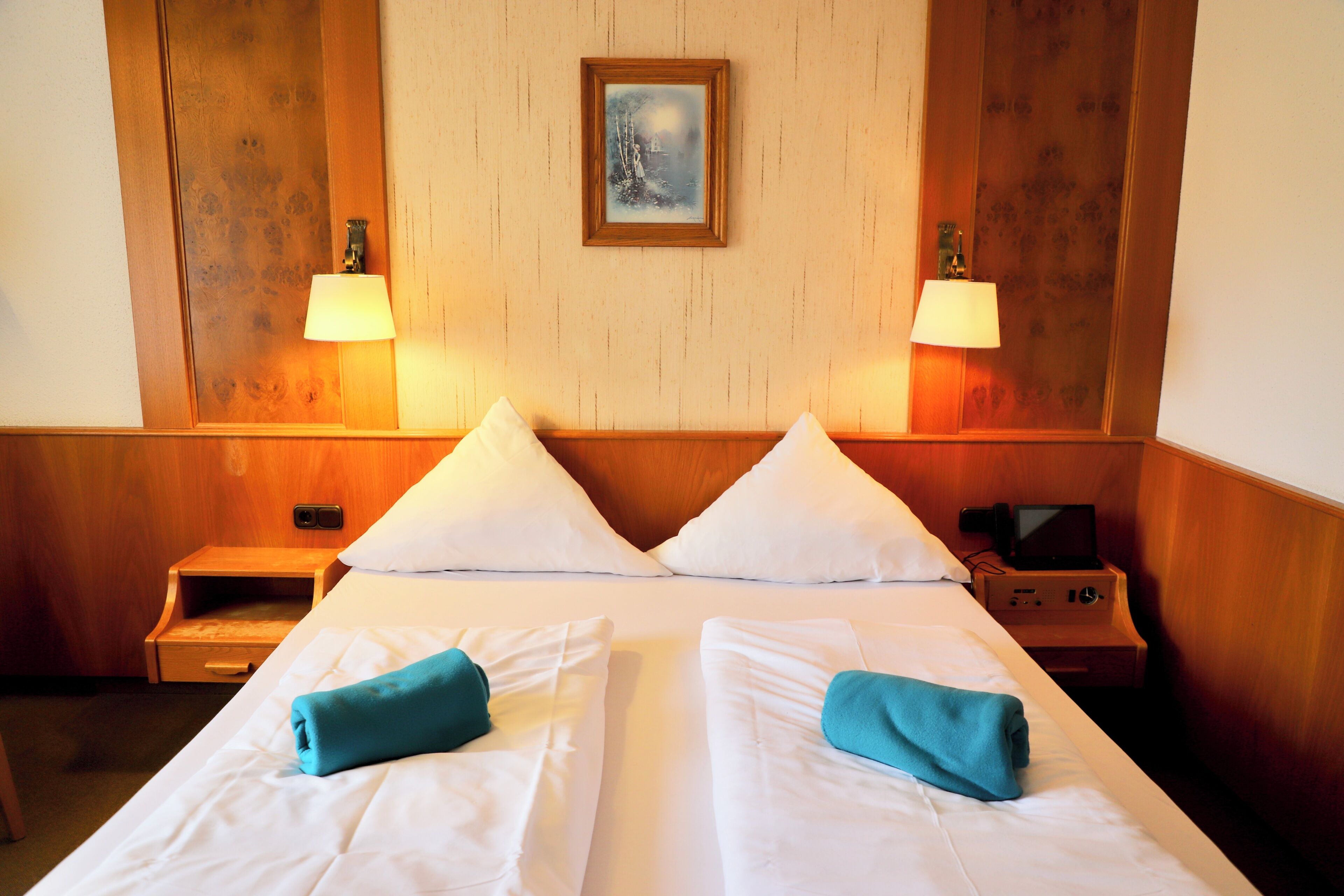 A hotel room with a double bed, two bedside lamps, a painting on the wall, and a small table with a lamp.