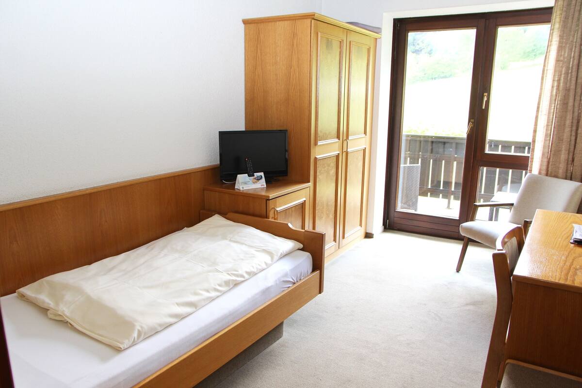Un lit simple avec une tête de lit en bois, un téléviseur posé sur un meuble en bois, une armoire en bois et un balcon avec une chaise.