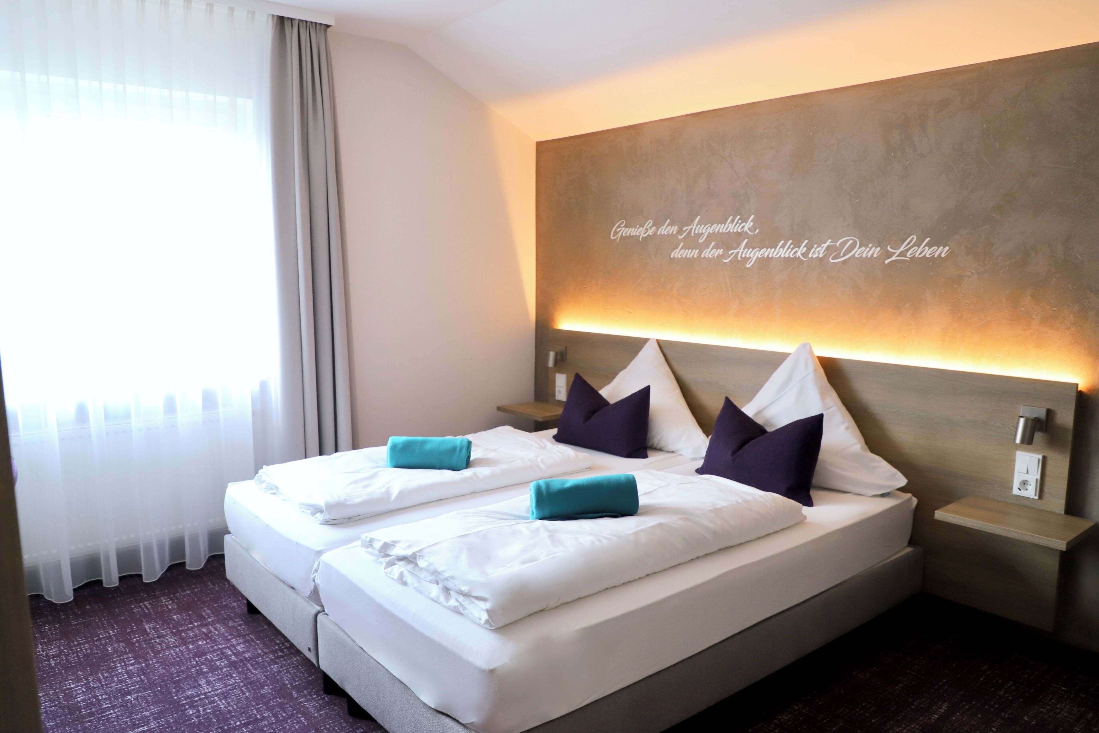 A hotel room with a double bed, two pillows, a nightstand, a wall-mounted light, and a window with curtains.