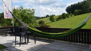 A hammock on a wooden deck with outdoor furniture, overlooking a green hillside.